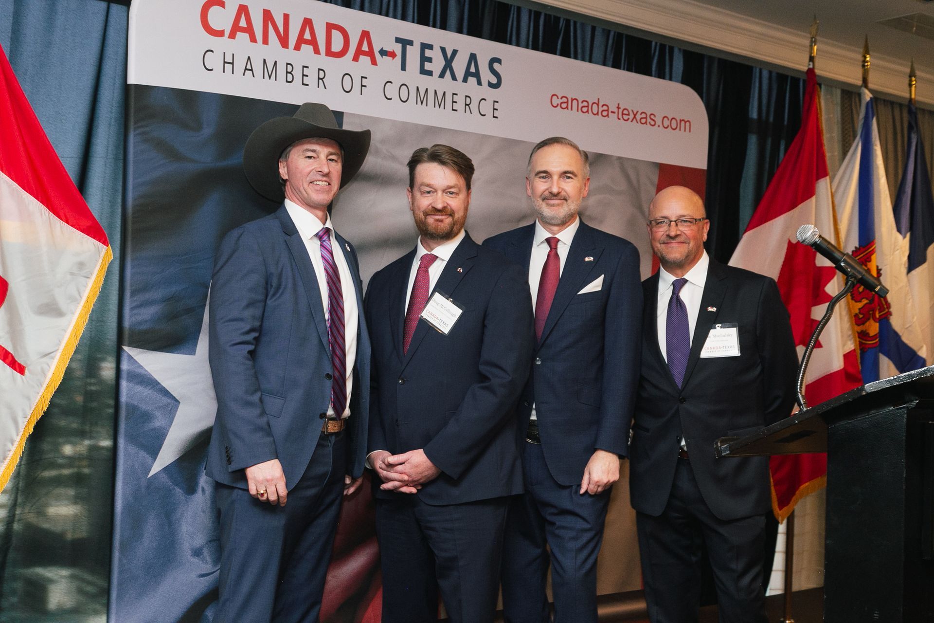 Four men in suits stand in a row before a Canada-Texas Chamber of Commerce backdrop and national flags.