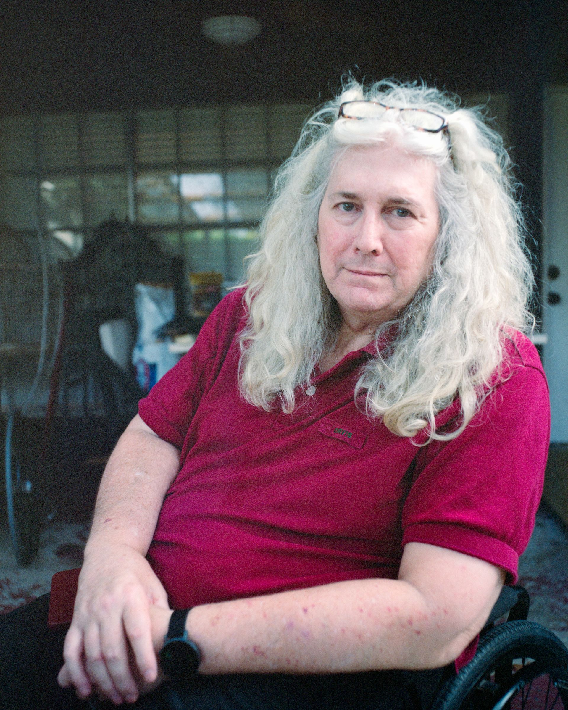 A person with long gray hair and glasses on their head wears a maroon polo shirt while seated in a wheelchair.