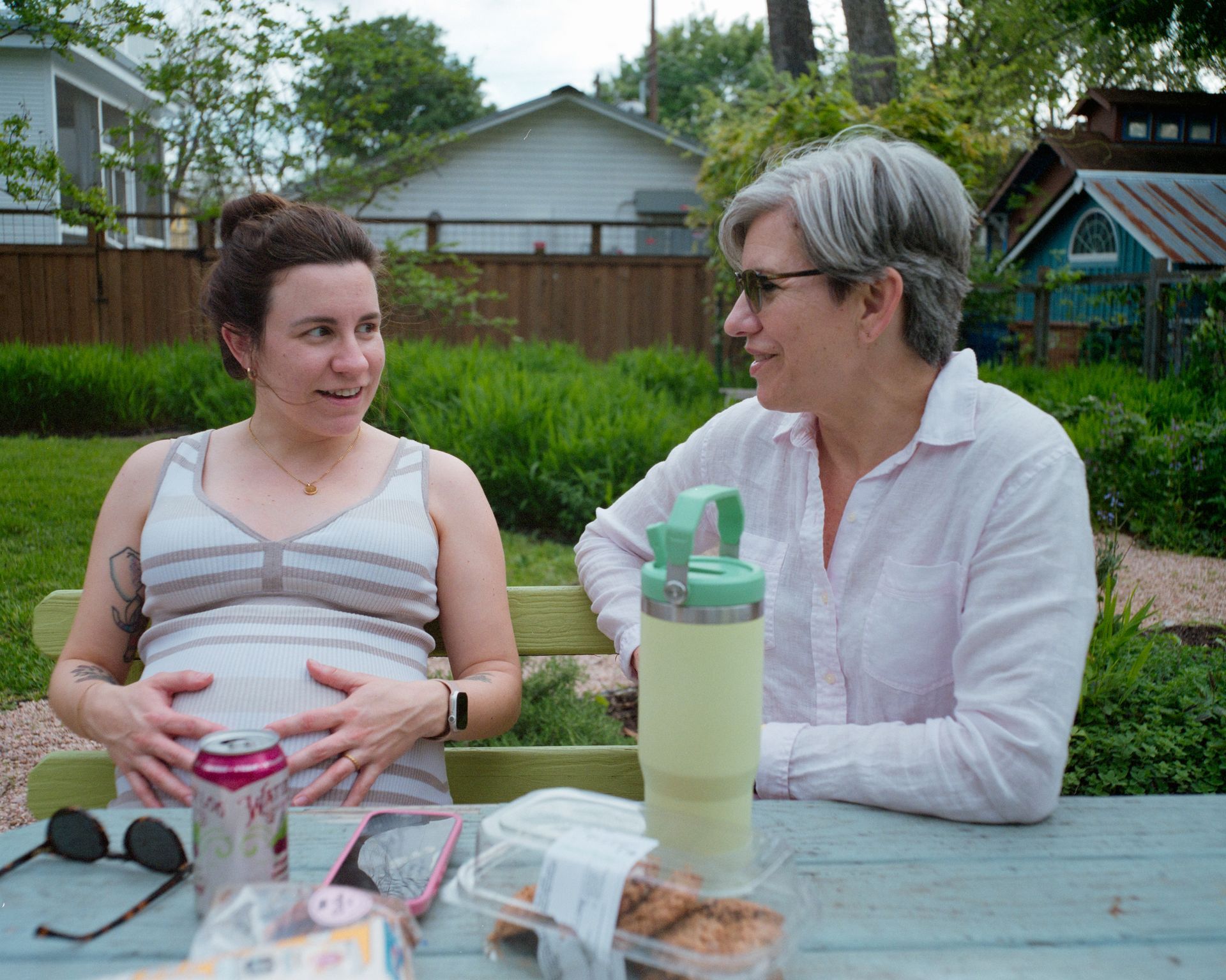 A pregnant person and an adult talk while sitting at an outdoor table in a backyard.