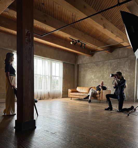 A woman knees down to photograph a woman standing opposite. A man sits on a couch next to the photographer and smiles at the subject.