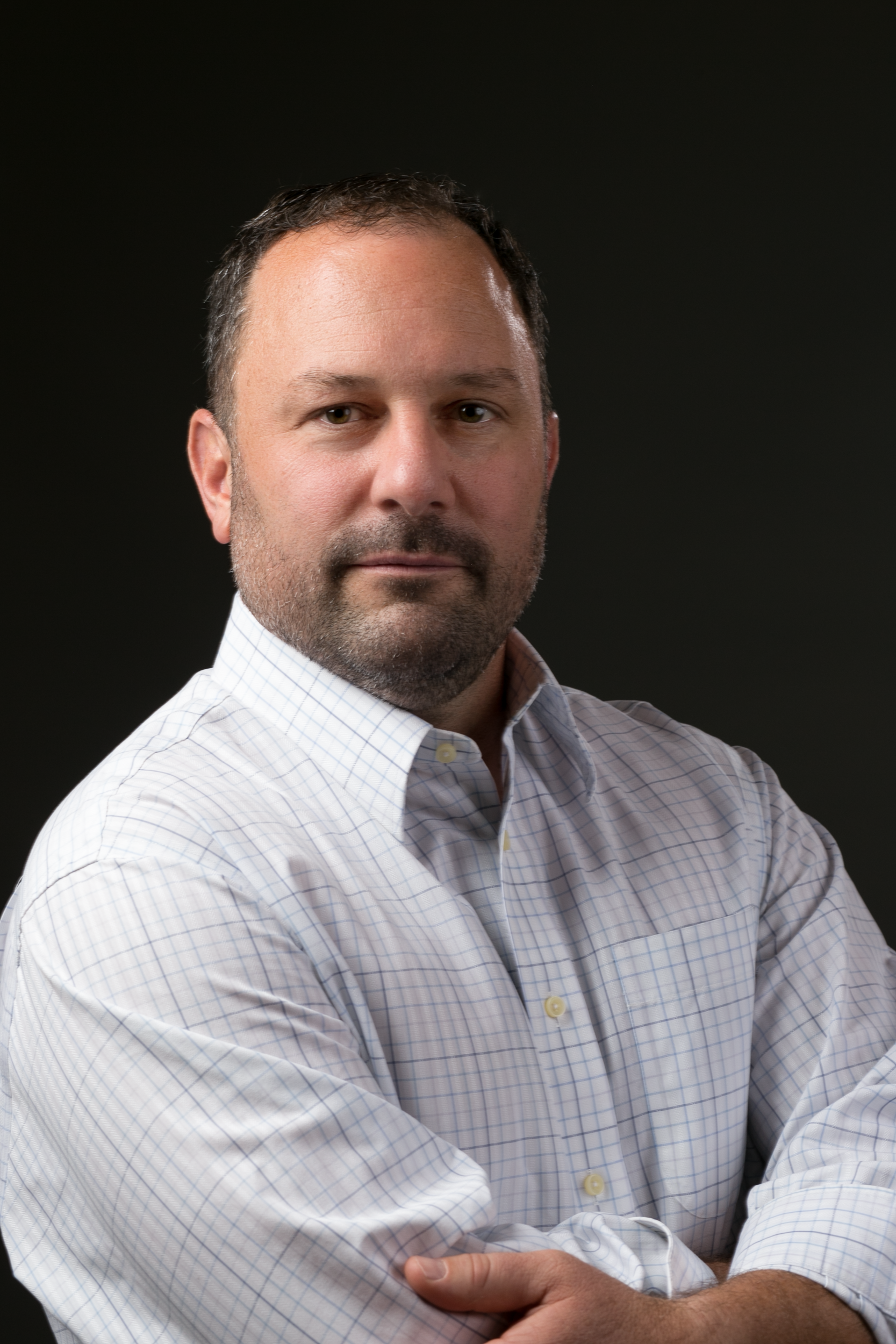 A man with a beard is wearing a white shirt and standing with his arms crossed.