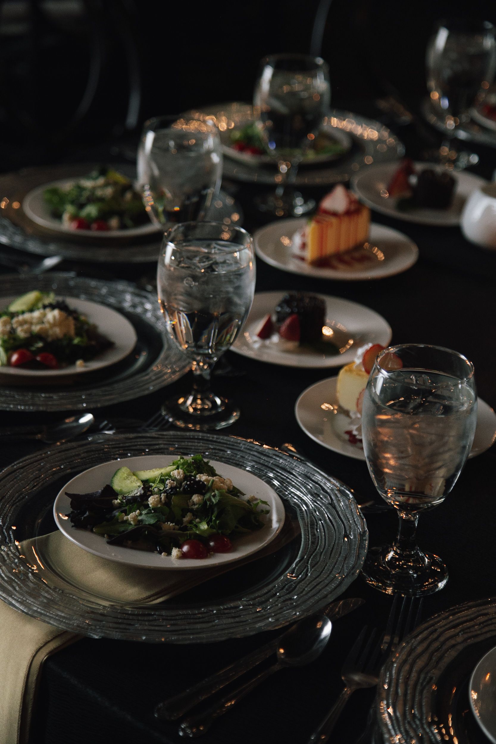 A dimly lit, formal dinner table set with elegant glassware, decorative chargers, and plates of salad and dessert.