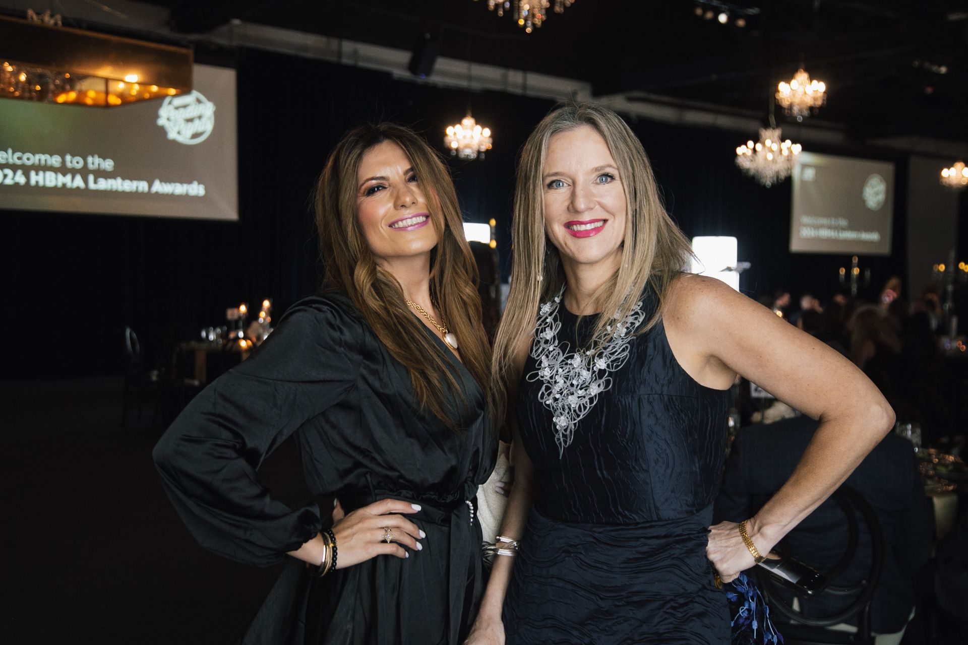 Two people smile at the camera in formal wear at the HBMA Lantern Awards ceremony, set in a dim banquet hall with chandeliers.