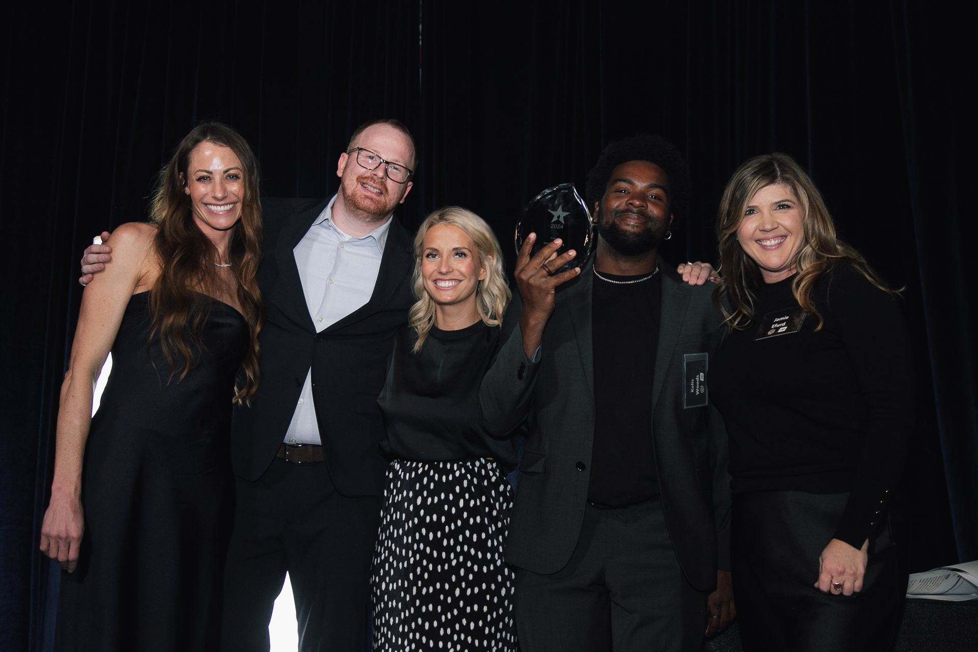 A group of five people stand in front of a black curtain, smiling as the person second from the right holds an award.