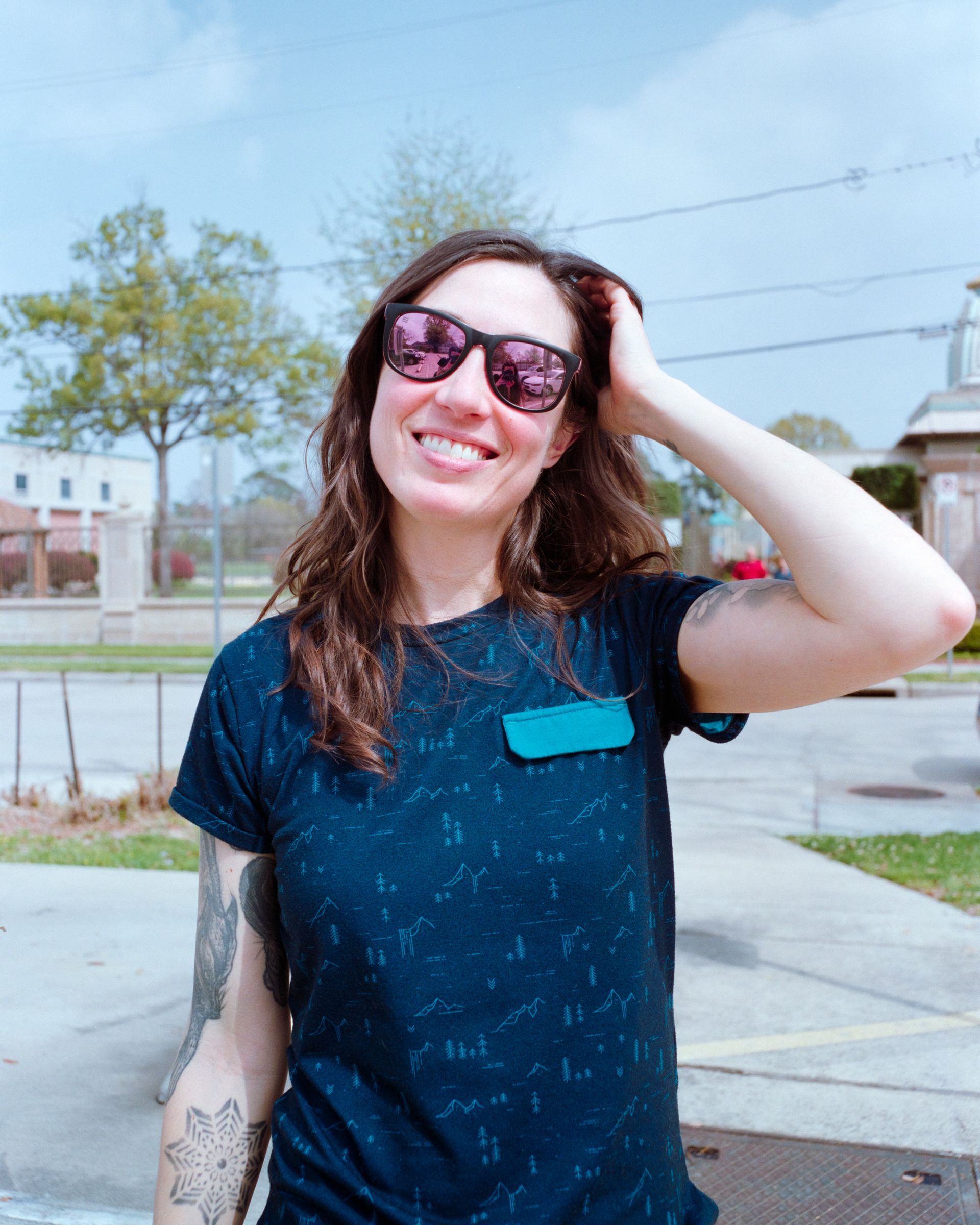 A smiling person wearing sunglasses and a patterned navy t-shirt stands outside with their hand touching their hair.