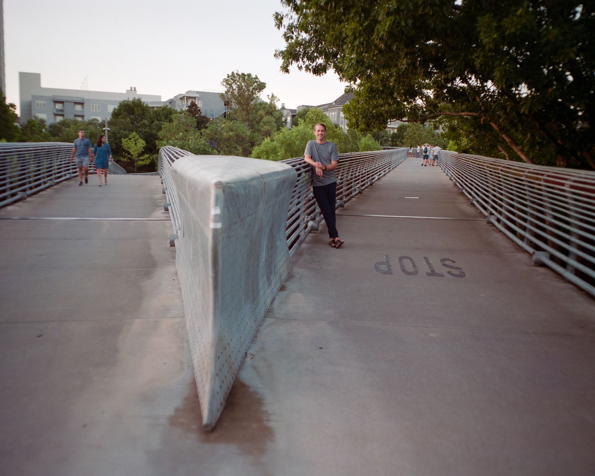 A person leans against a metal barrier on a pedestrian bridge, with others walking in the distance.