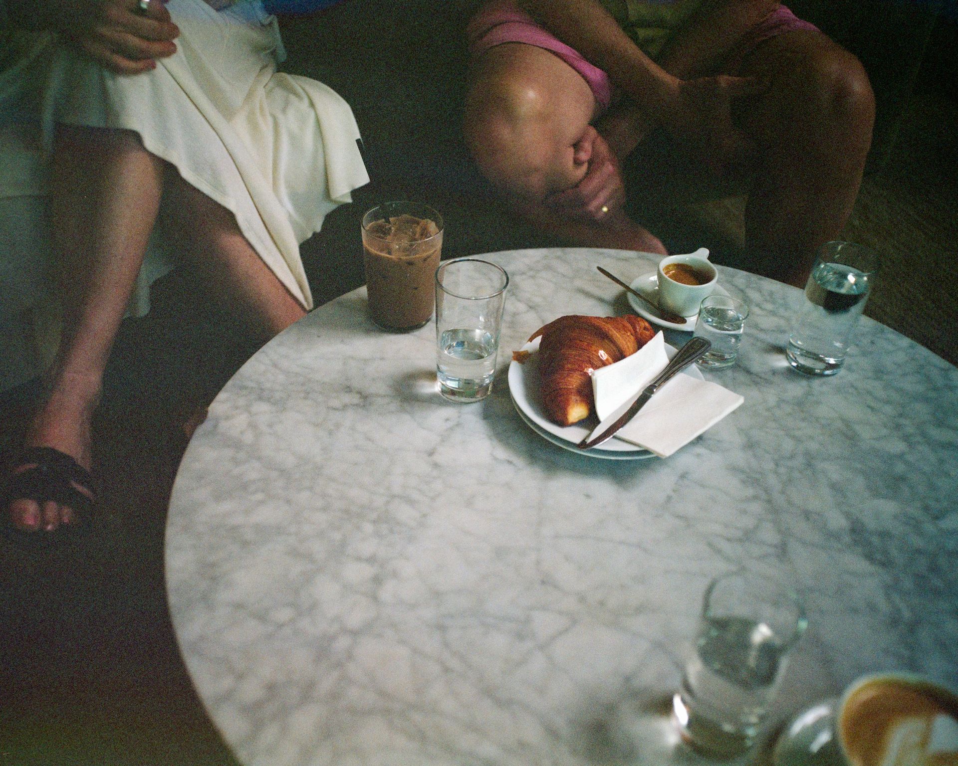 A marble café table holds coffee, water, and a croissant, with two people seated behind it.