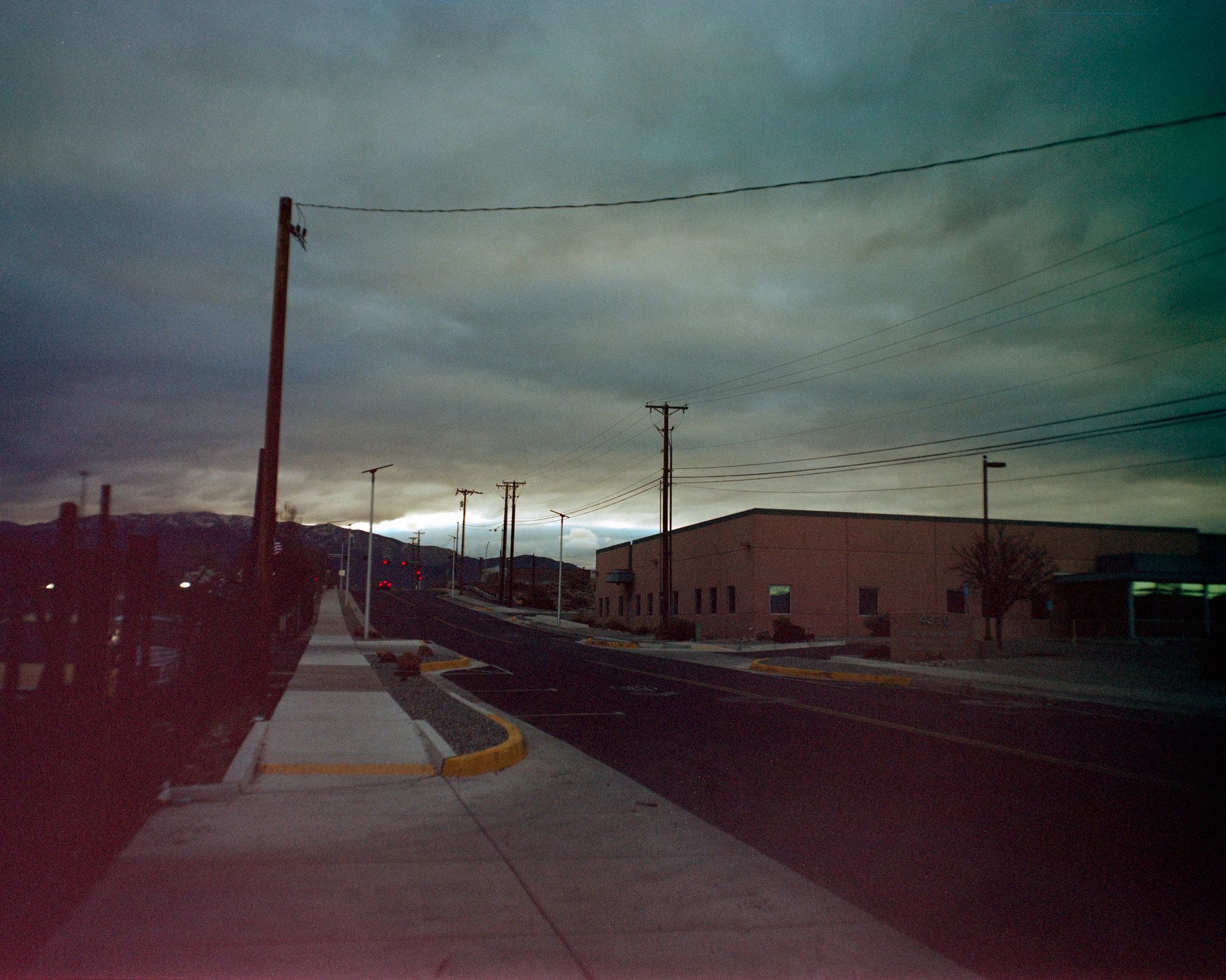 A paved sidewalk leads toward a road beside a building under a dark, cloudy sky at dusk.