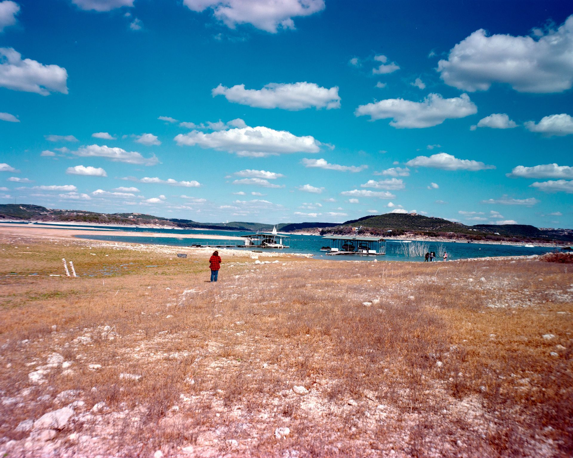 A person in a red jacket stands on a dry, grassy shore overlooking a blue lake with docked boats under a cloudy sky.