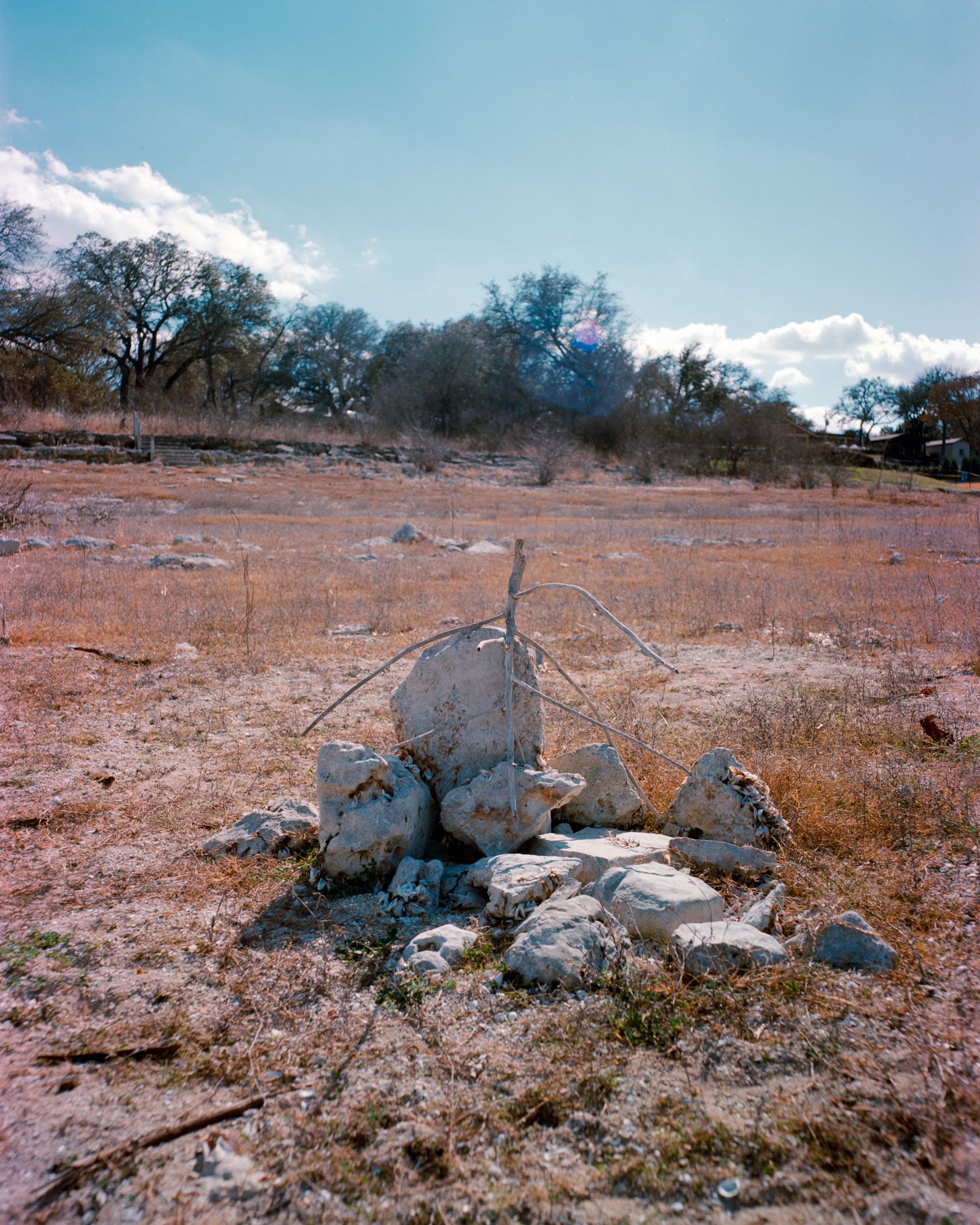 A small pile of light-colored rocks topped with a thin wooden cross in a dry, sparse field under a bright blue sky.