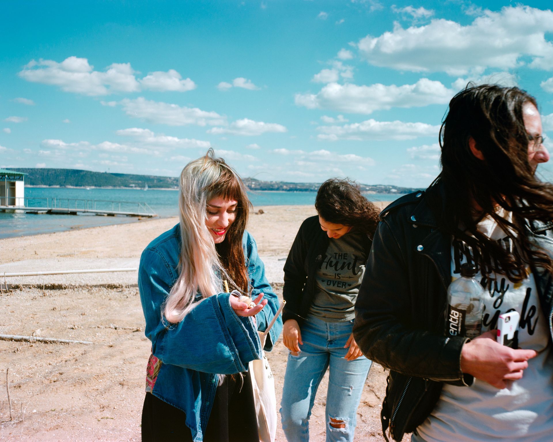 Three friends walk together on a sunny beach, smiling and looking down while talking.