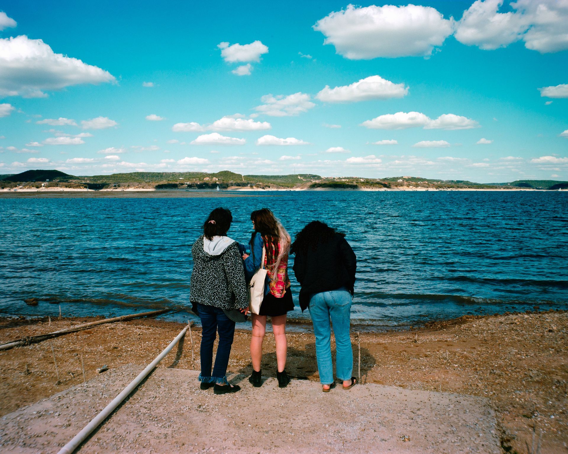Three people stand on a shore, backs to the camera, looking out over a blue lake under a sky with scattered white clouds.