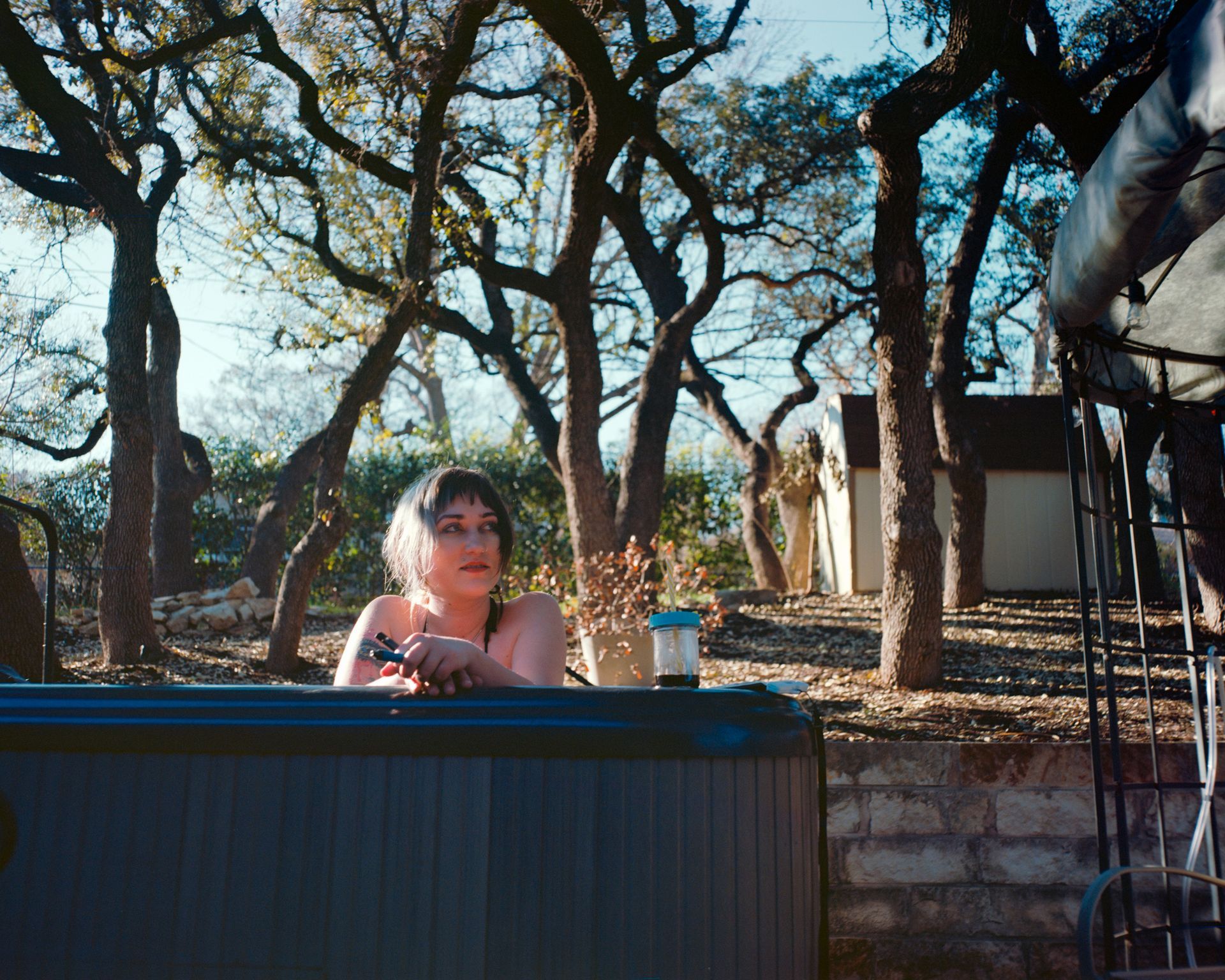 A person rests in a hot tub outdoors among trees, looking toward a small building in a sunny, wooded backyard setting.