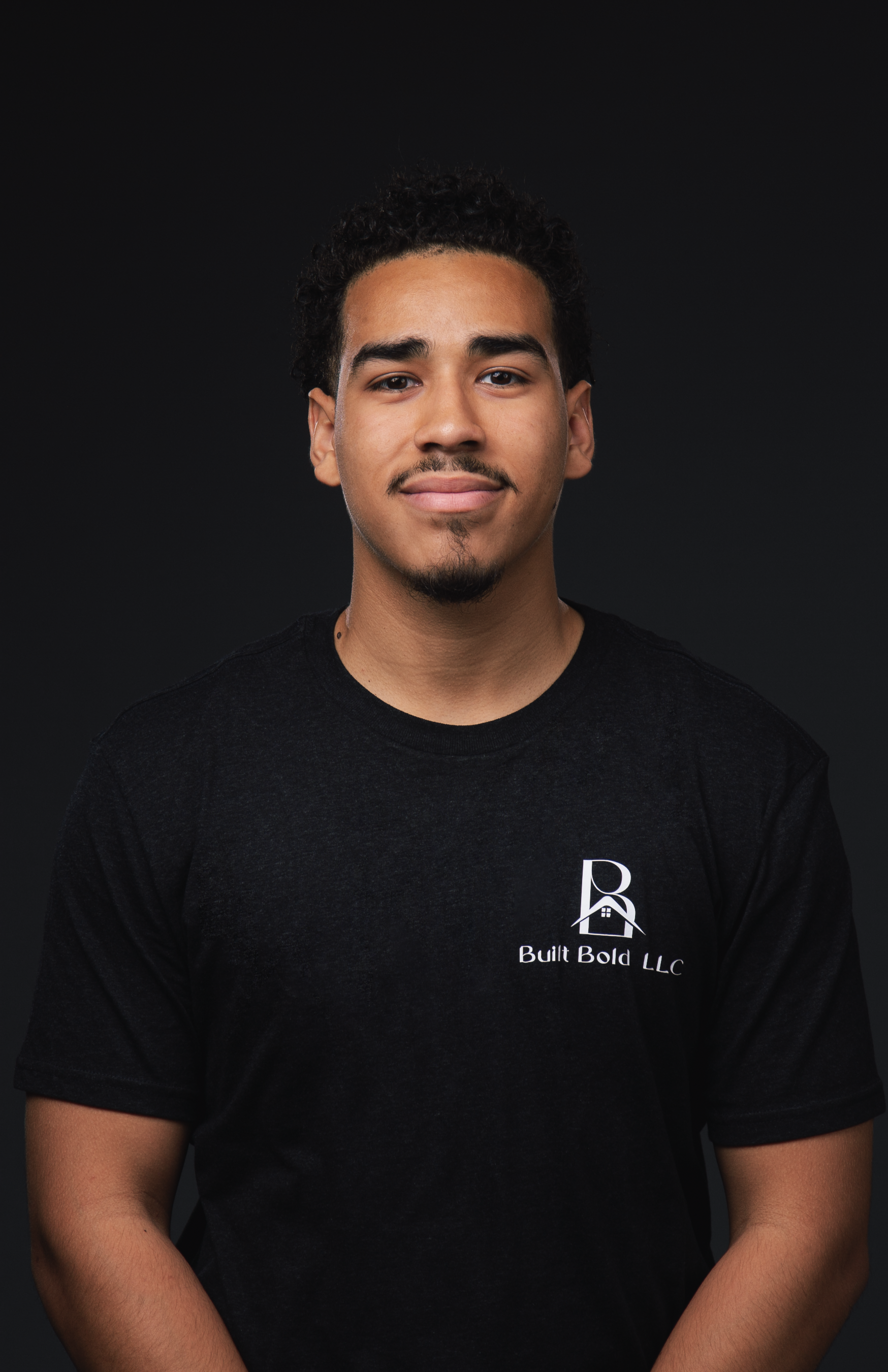 A man with a beard is wearing a white shirt and standing with his arms crossed.