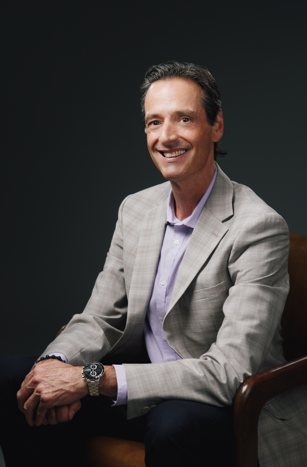 A man in a suit is sitting in a chair with his hands folded and smiling.