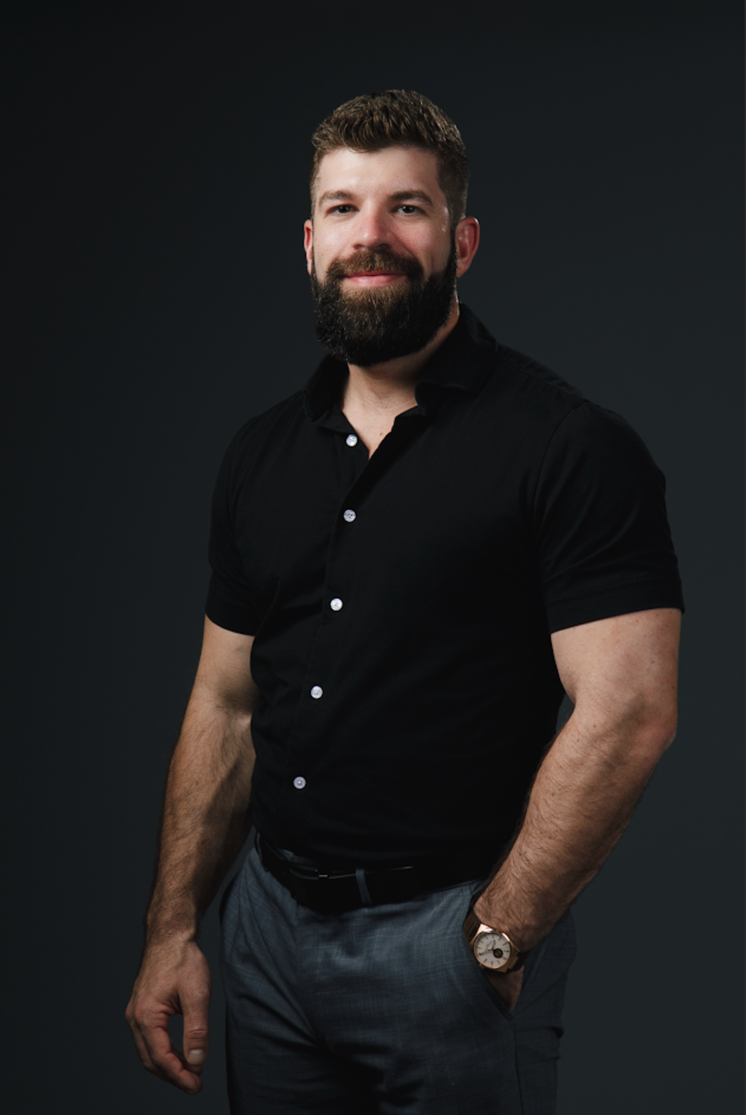 A man with a beard is wearing a black shirt and gray pants.