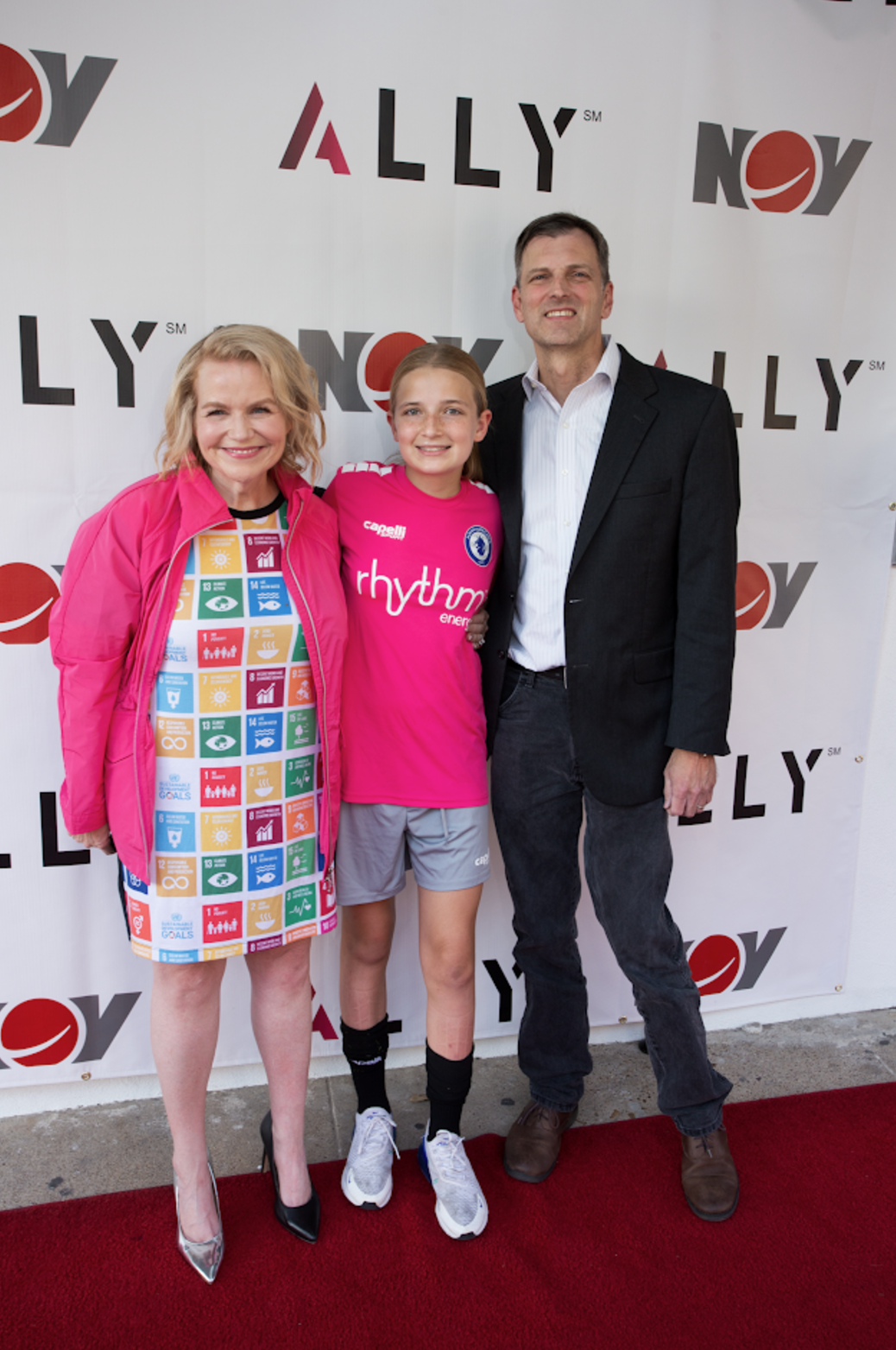 A woman in a pink jacket is standing next to a man and a girl on a red carpet.