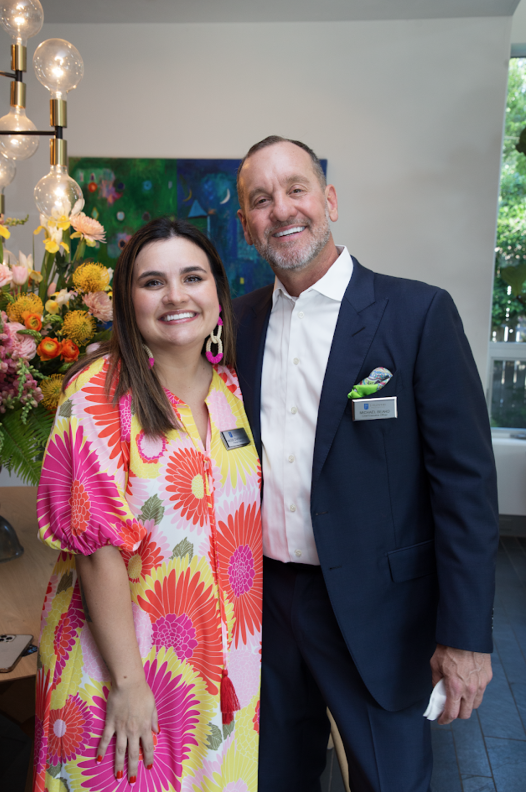 A woman in a vibrant floral dress and a man in a navy suit and white shirt smile together in a room with flowers.