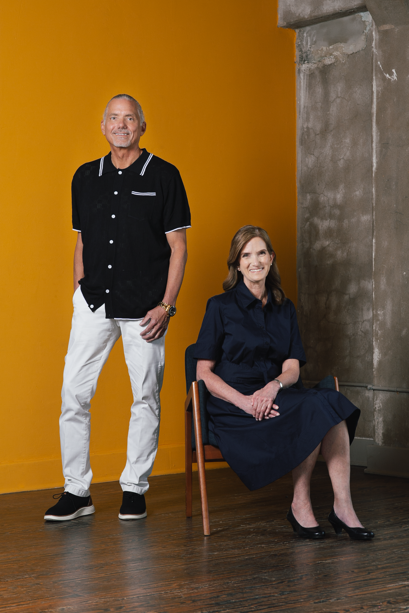 A formal portrait photography of a man standing and a woman sitting in a chair. They are in front of a yellow wall that juts up against a concrete wall. The floor is made of hardwood.