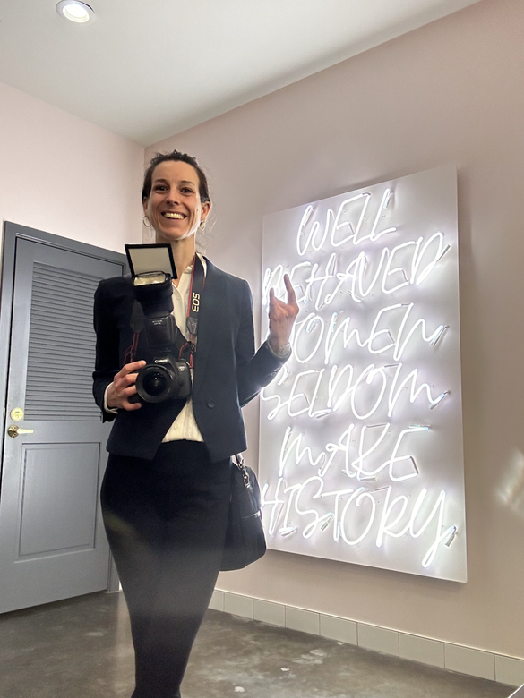 A woman is standing in front of a neon sign holding a camera.