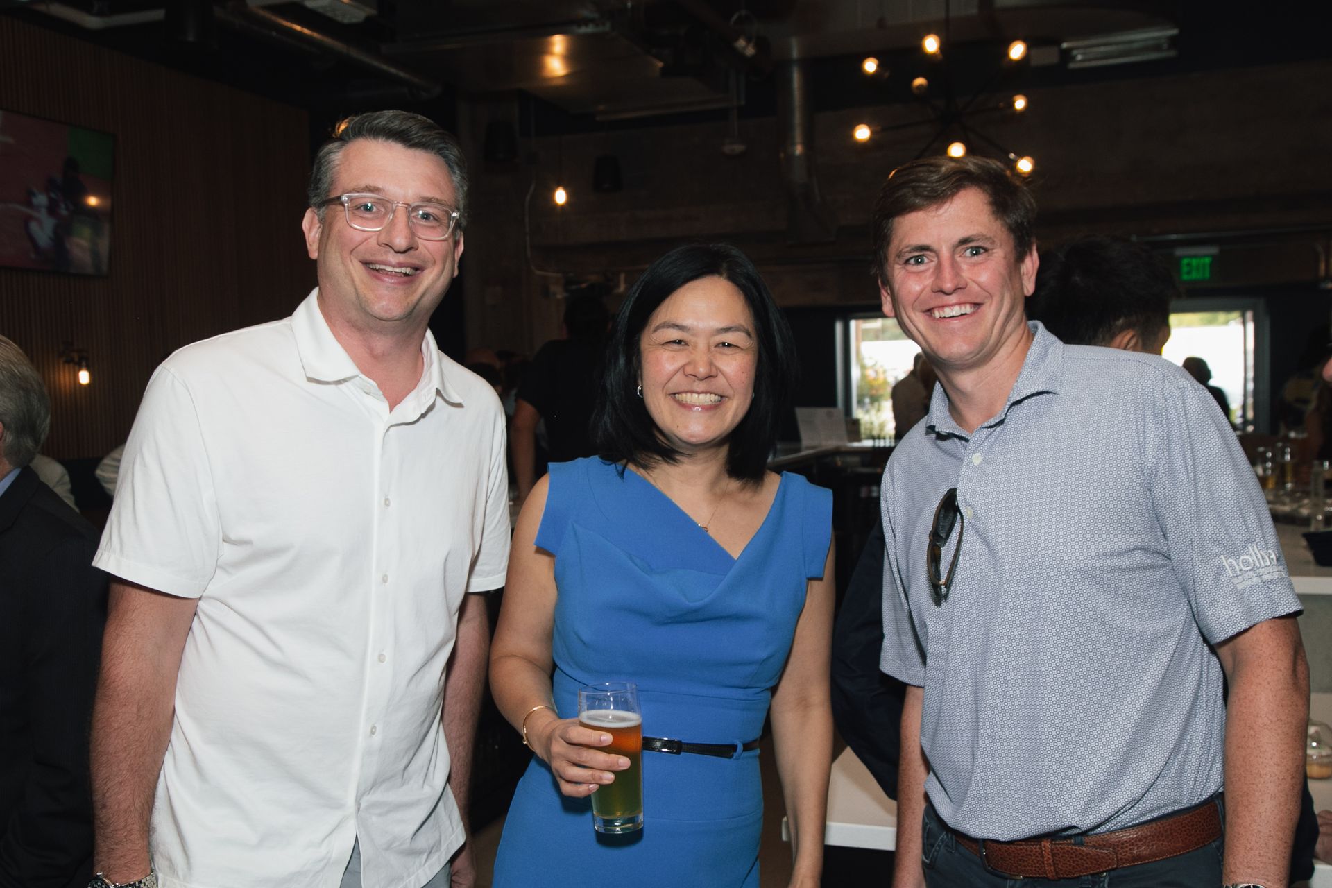 Three people smiling while standing together at an indoor social event; the person in the middle holds a glass of beer.