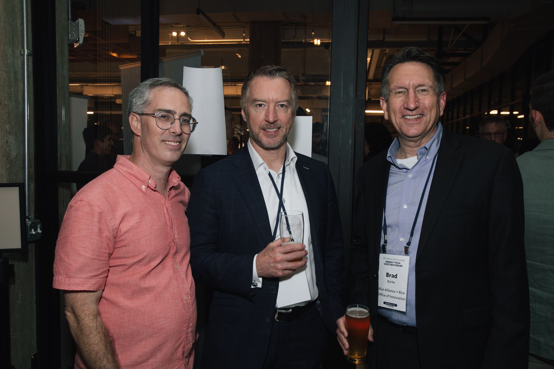 Three people smile in a casual indoor setting, one in a pink shirt and two in business attire with conference badges.