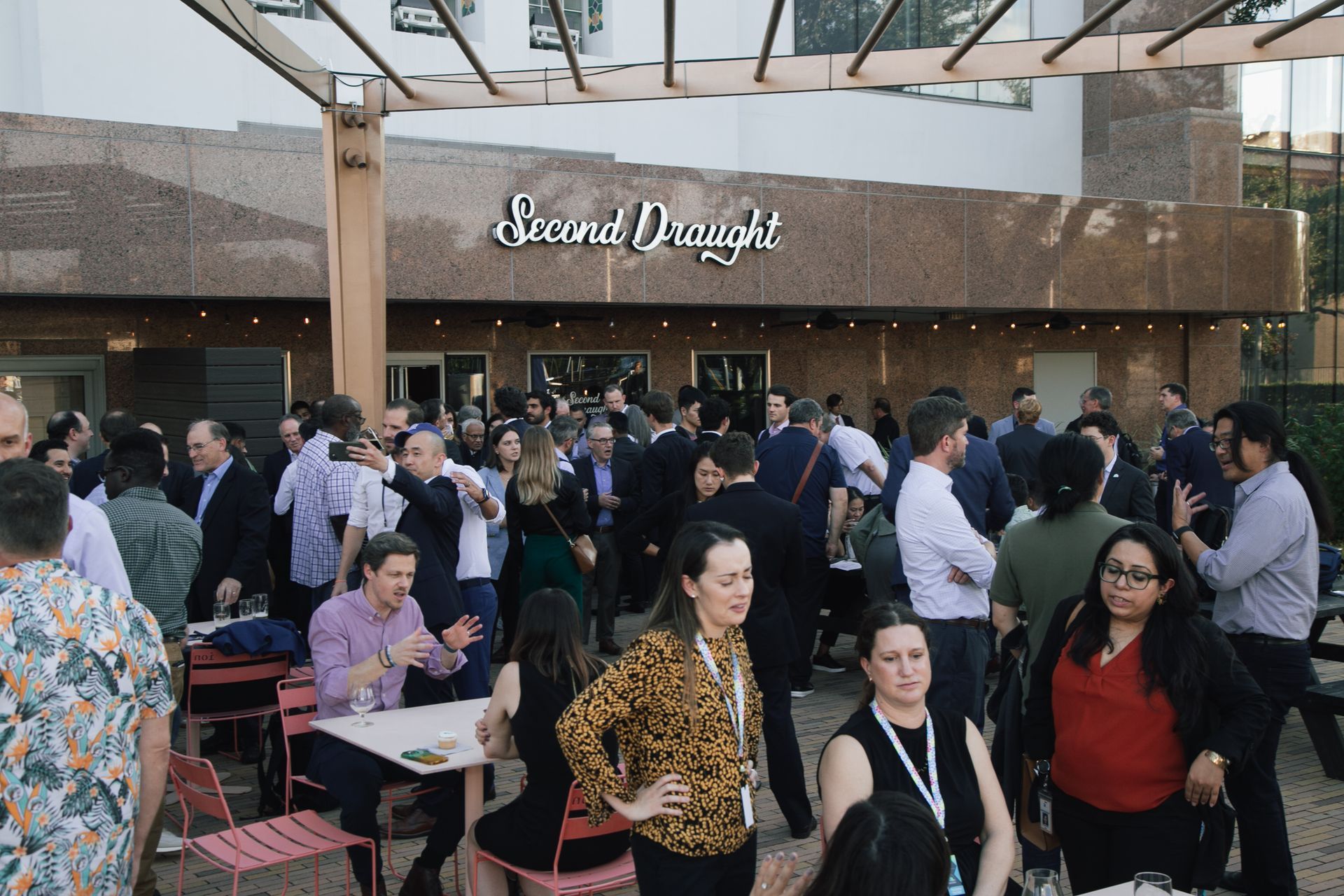 A crowded outdoor social gathering at a restaurant called Second Draught, with people standing and sitting at tables.