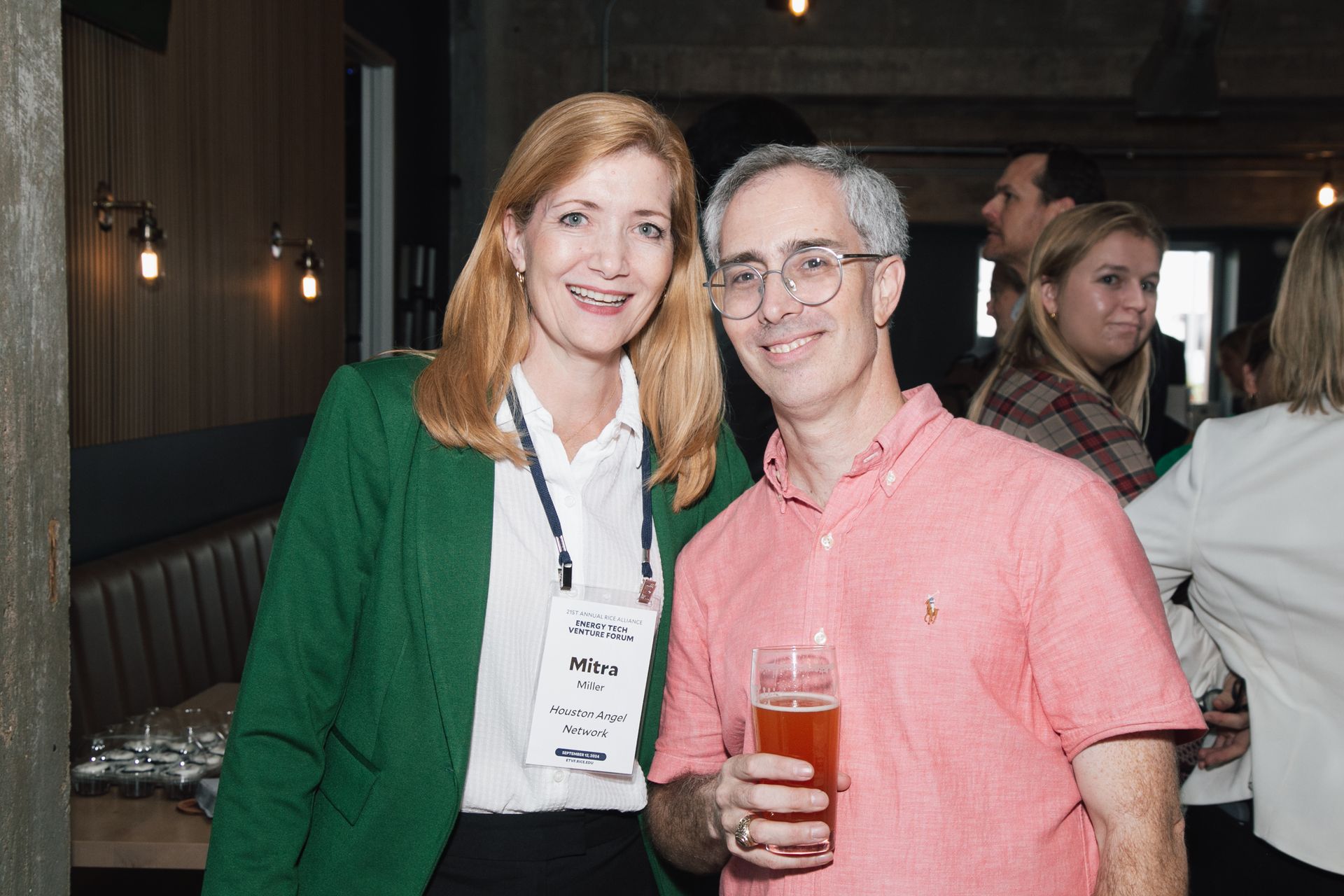Two colleagues smile while standing at an indoor social event; one wears a green blazer and the other a pink polo shirt.