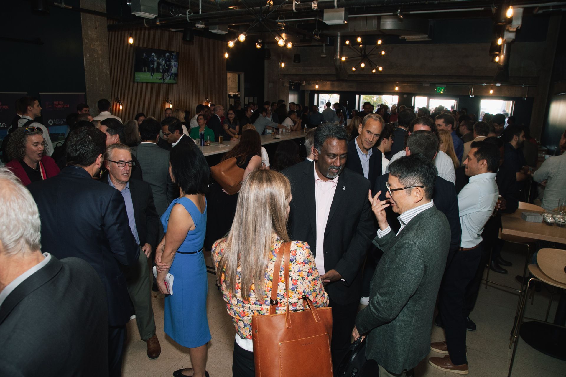 A crowded indoor social event with people in business-casual attire conversing in a dimly lit, contemporary bar setting.