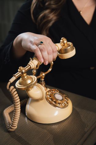 A person wearing a ring holds the handset of a vintage, cream-colored rotary telephone on a dark surface.