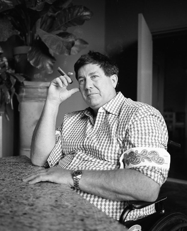 A man in a checkered shirt holding a cigar, sitting at a counter with his sleeves rolled up to show the deorative edge.