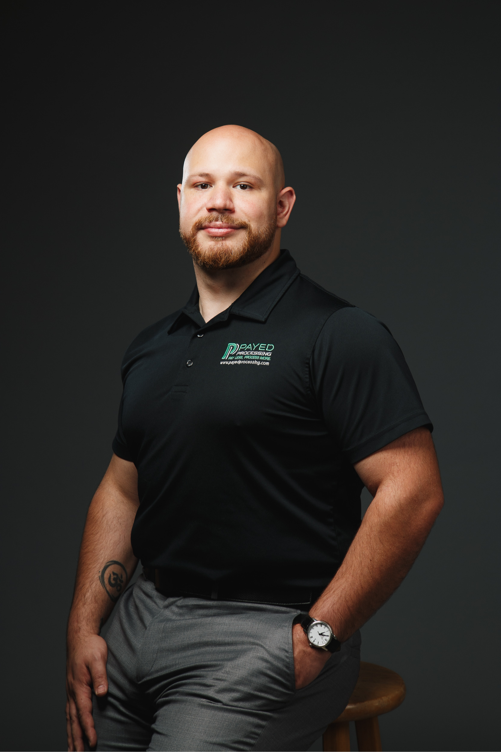 Man in black polo, gray pants, hand in pocket, looking at the camera with a slight smile.