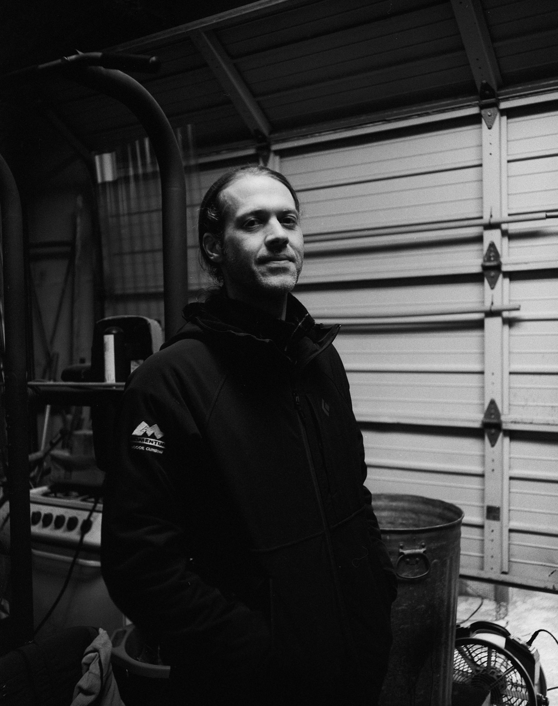 A person in a dark jacket stands in a garage, looking toward the camera with a neutral expression in black and white.