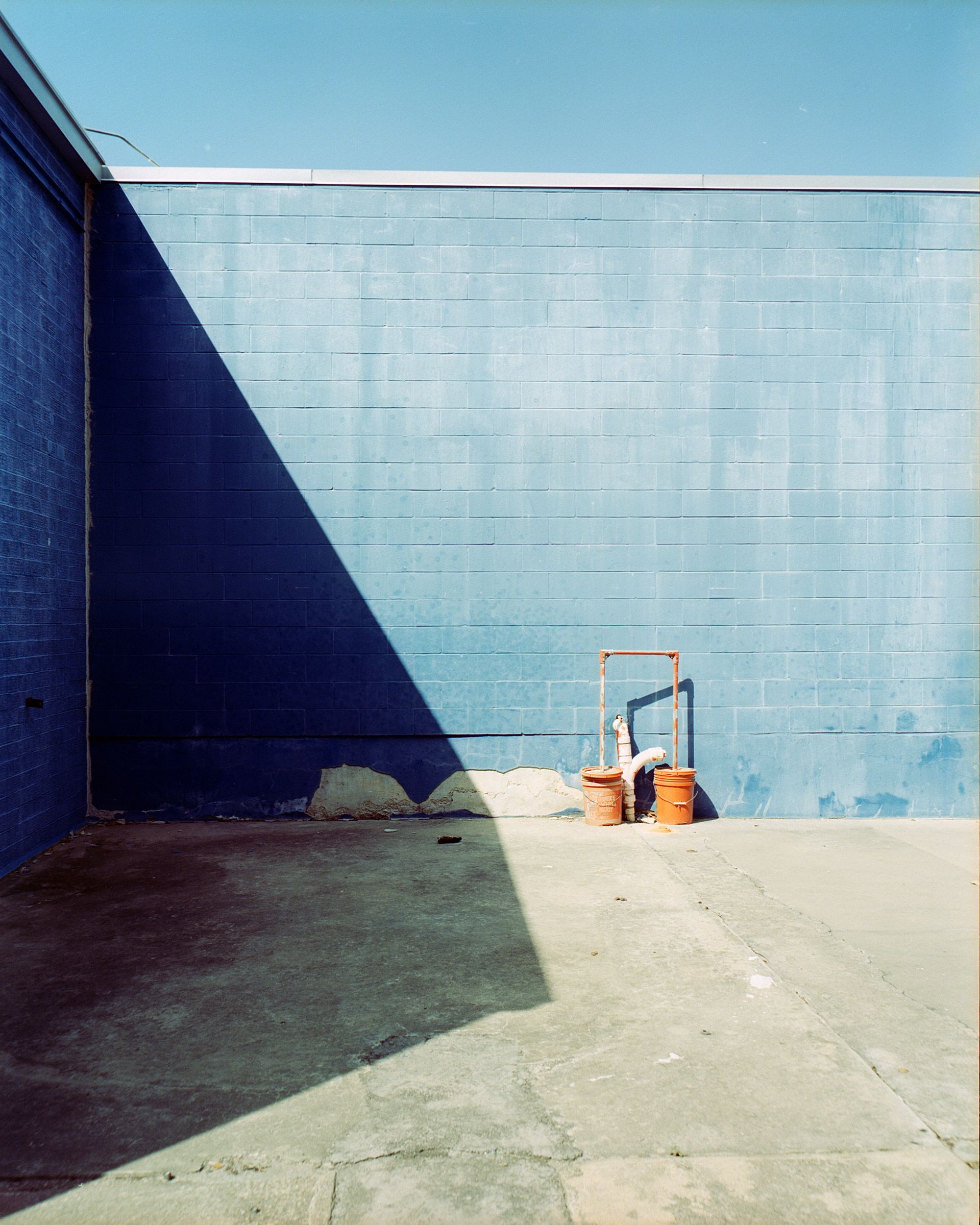 A long, triangular shadow falls across a blue cinder block wall, with two orange buckets sitting on the pavement below.