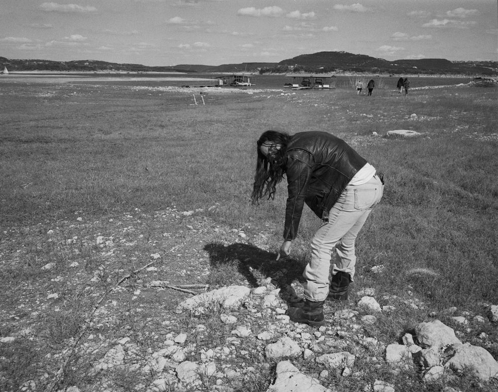 A person with long hair wears a leather jacket and jeans while bending down to examine rocky ground in an open field.