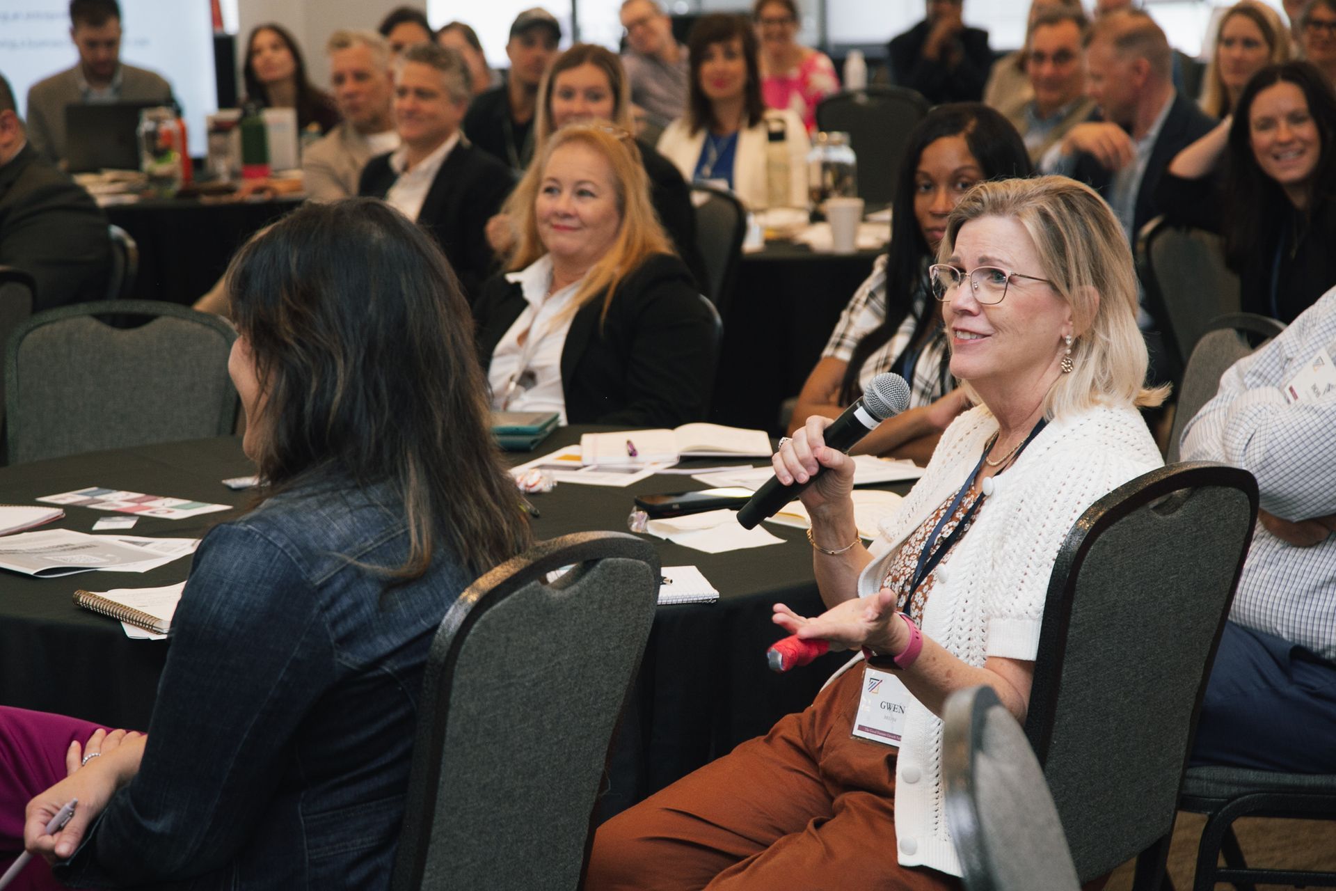 Attendees at a professional conference sit at round tables, with one individual speaking into a microphone.