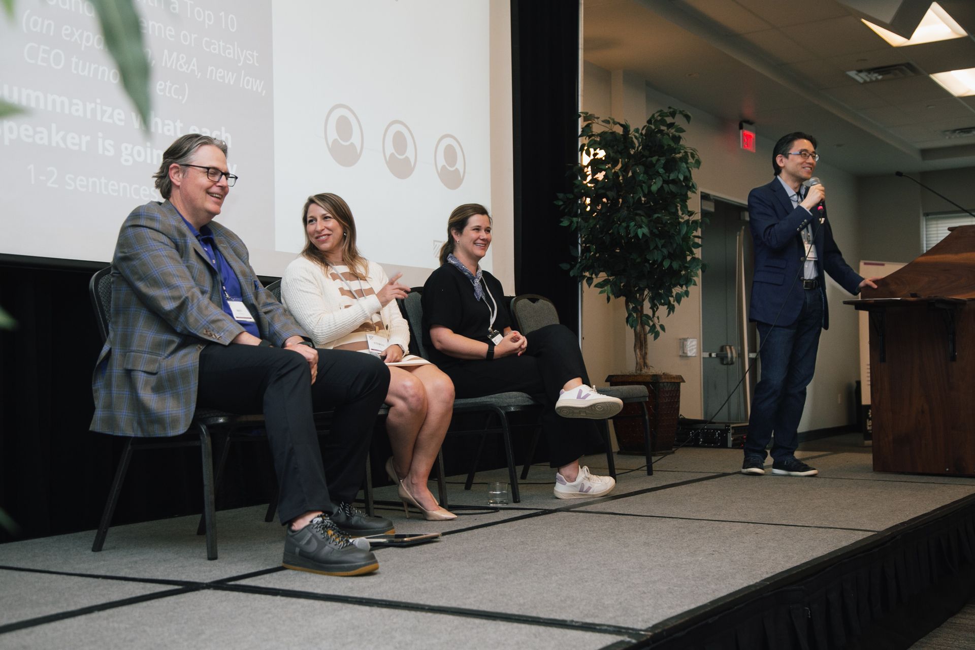 A panel of four people on a stage: three seated in chairs and one standing at a lectern speaking into a microphone.