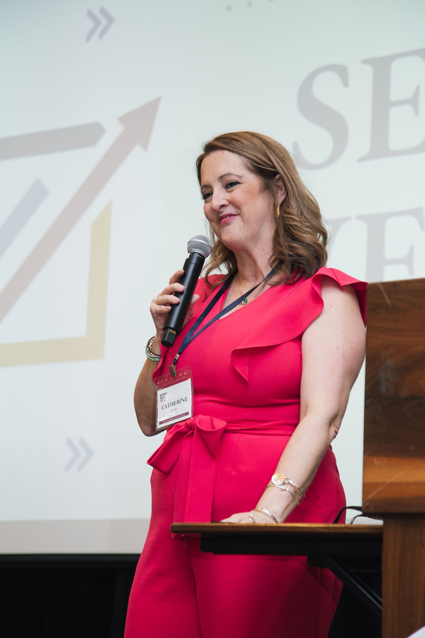 A smiling presenter in a bright pink jumpsuit speaks into a microphone while standing at a podium.