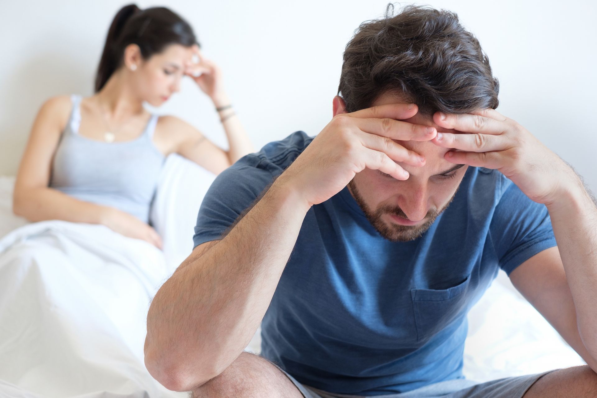 Couple sitting apart on a bed, both with hands on their heads, looking distressed.