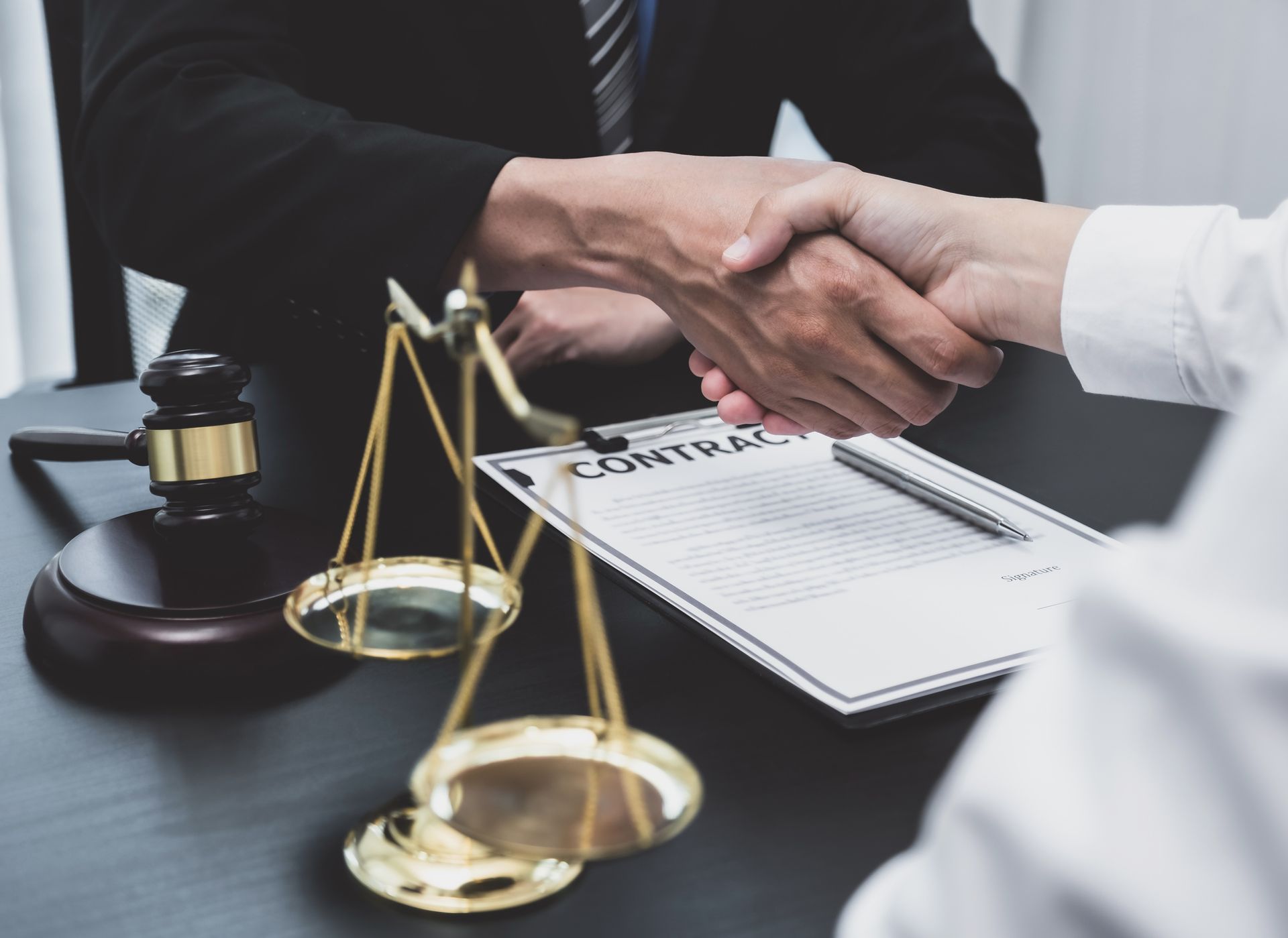 Two people shaking hands over a contract and scales of justice, gavel nearby. Two people shaking hands over a contract and scales of justice, gavel nearby.