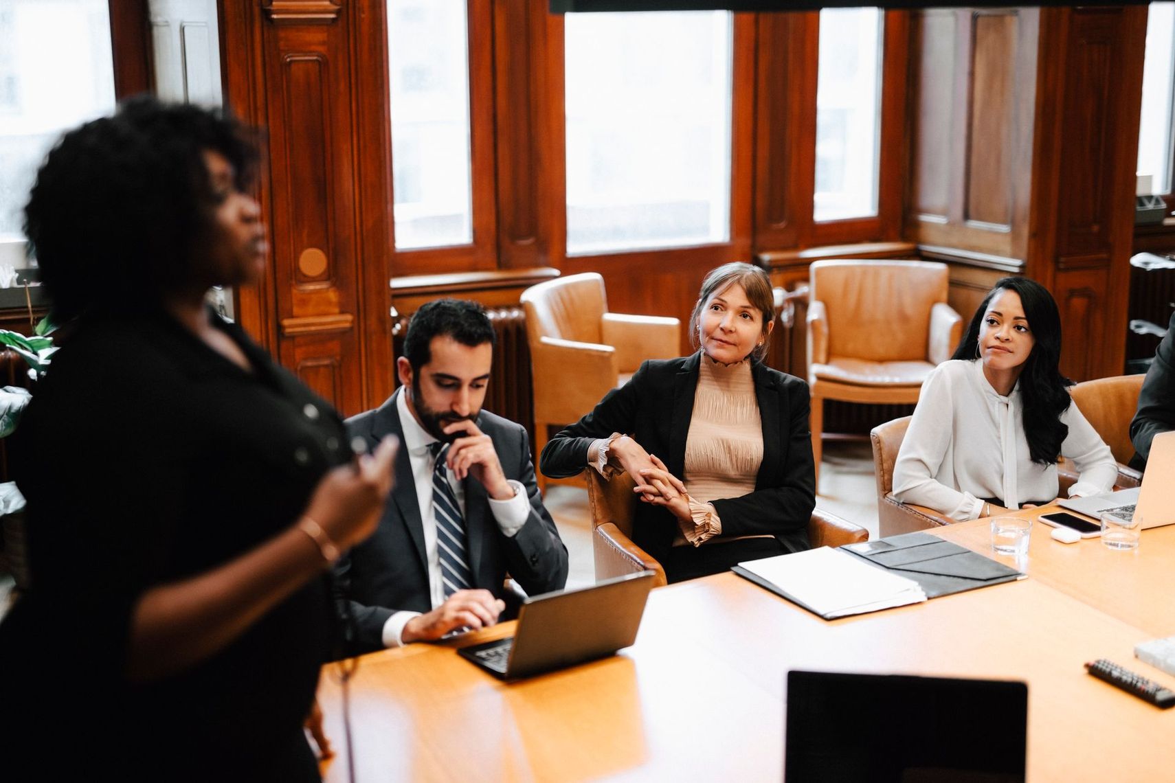 A woman giving a presentation to a diverse group seated around a conference table in a wood-paneled room.
