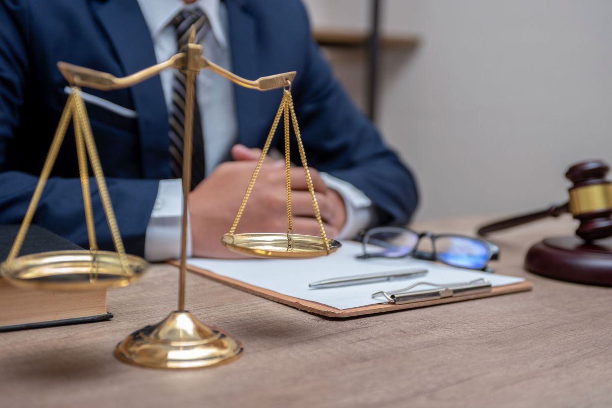 Scales of justice, legal documents, and gavel on a desk with a person in a suit, suggesting legal counsel.