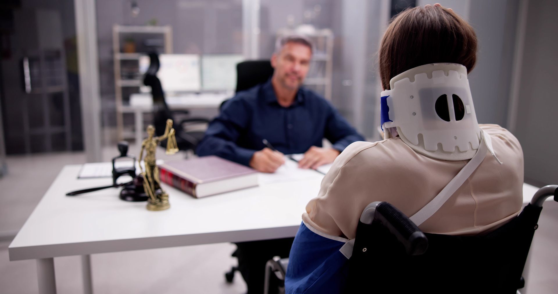 Person in a neck brace and cast in a wheelchair consulting with a lawyer at a desk. Person in a neck brace and cast in a wheelchair consulting with a lawyer at a desk.