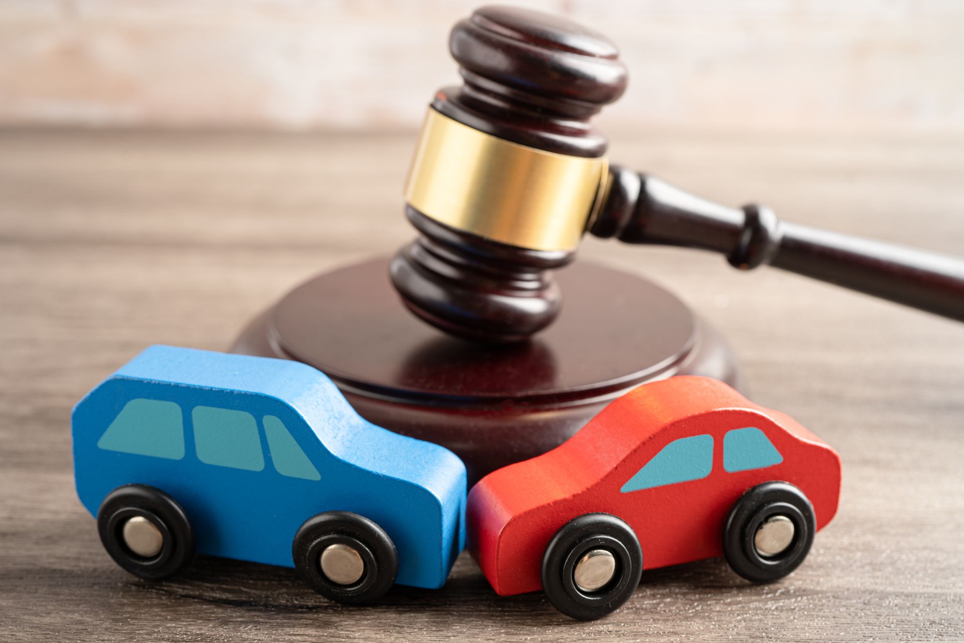 Lawyer and client review documents at a desk in an office. Gavel and scale visible. Blue and red toy cars with gavel on wooden surface, implying a car accident claim.