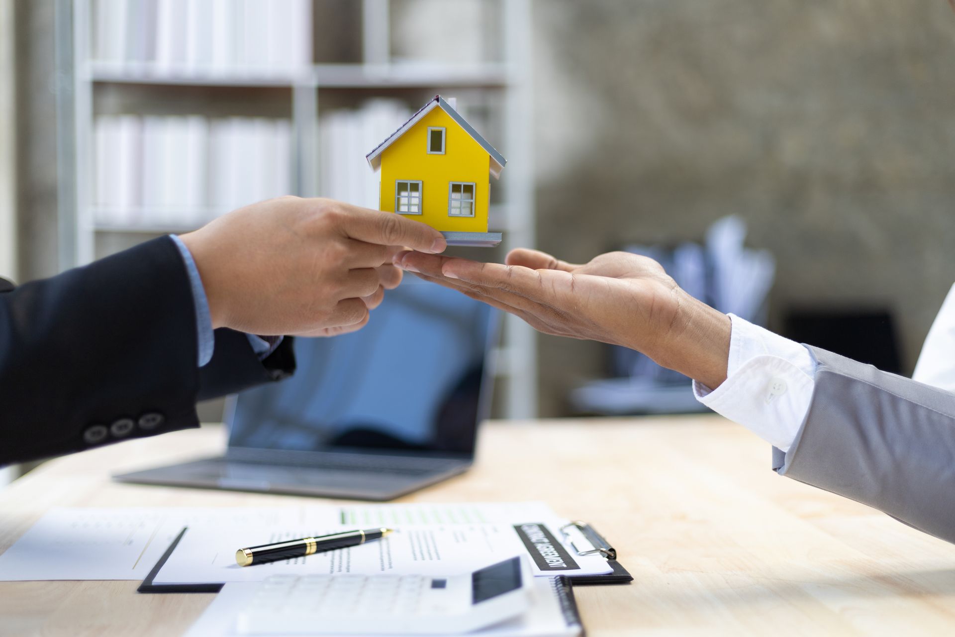 Two hands passing a yellow model house over a table with documents, pen, and laptop. Two hands passing a yellow model house over a table with documents, pen, and laptop.