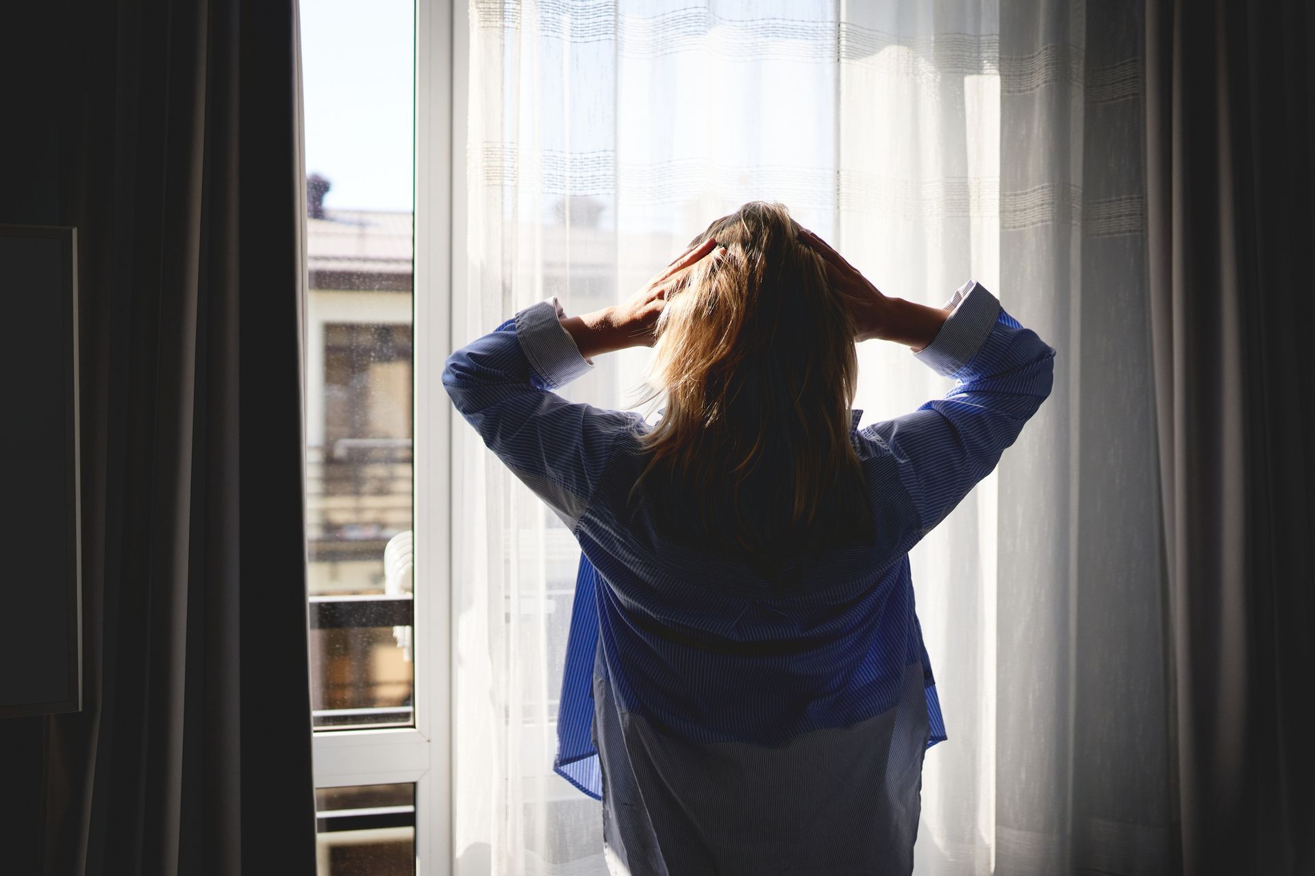 Woman standing in front of a window with hands on head, lit by sunlight. Woman standing in front of a window with hands on head, lit by sunlight.