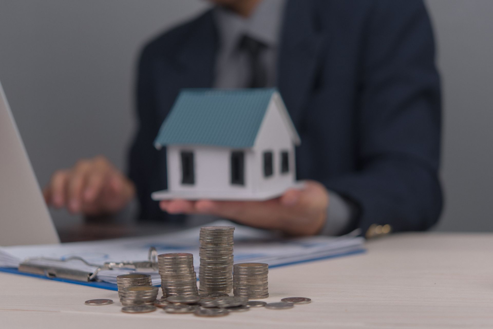 Person holding a model house, stacks of coins on a table with a laptop and documents, symbolizing real estate investment. Person holding a model house, stacks of coins on a table with a laptop and documents, symbolizing real estate investment.