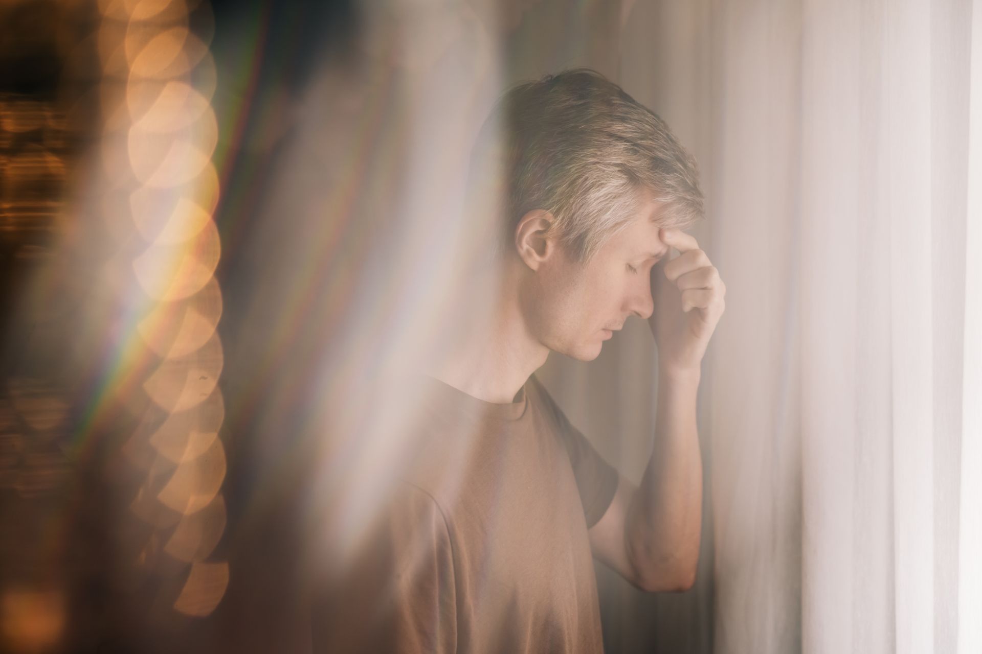 Man with hand on forehead, looking down; blurred, soft-focus, near window. Man with hand on forehead, looking down; blurred, soft-focus, near window.