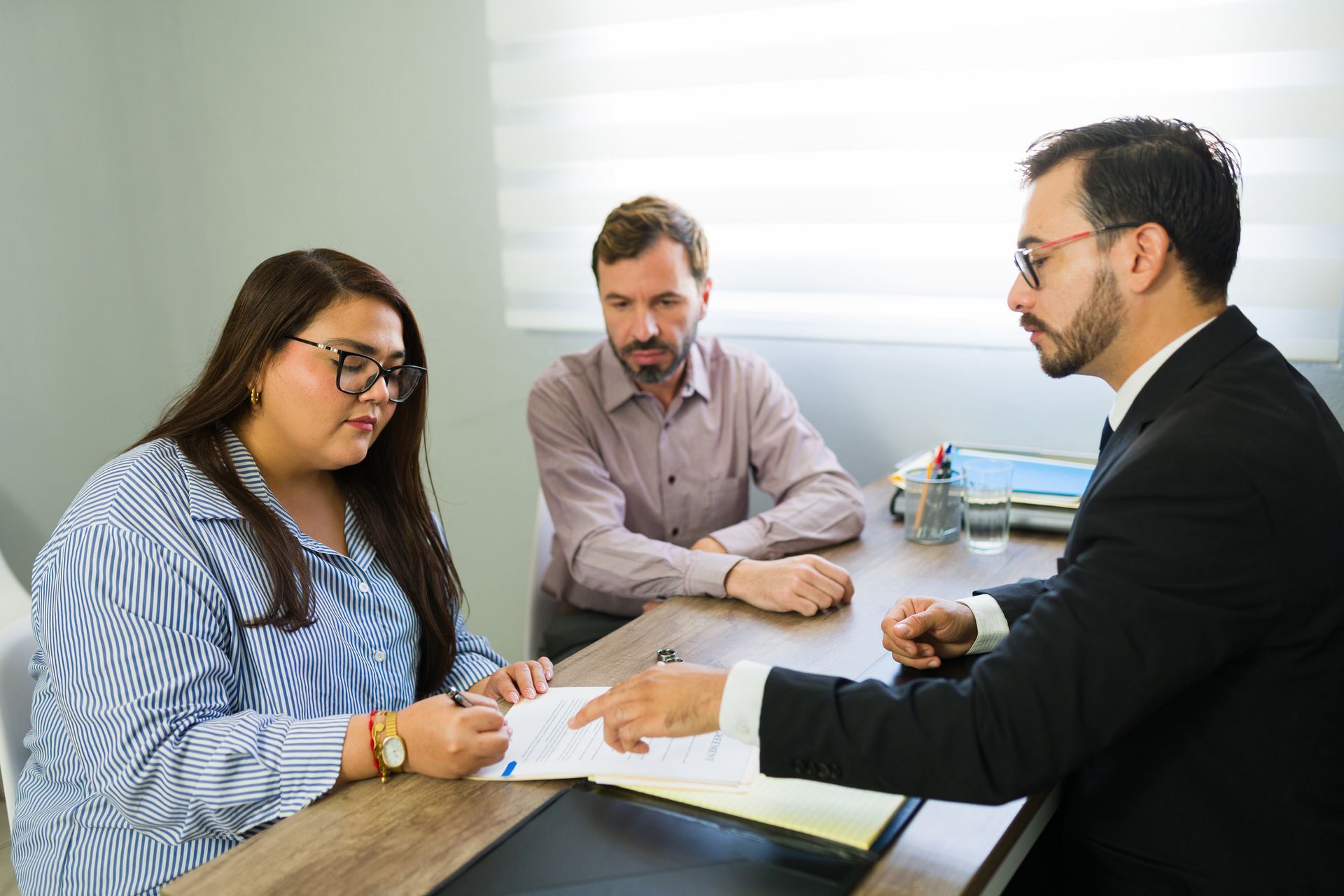 Woman signing document at a table, man looks on, lawyer points; office setting. Woman signing document at a table, man looks on, lawyer points; office setting.