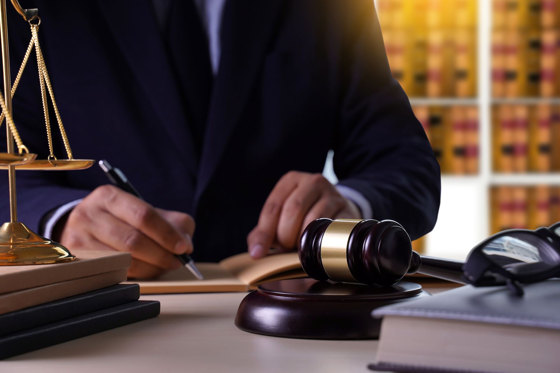 Person in suit writing, scales of justice, gavel, and law books on a desk. Person in suit writing, scales of justice, gavel, and law books on a desk.